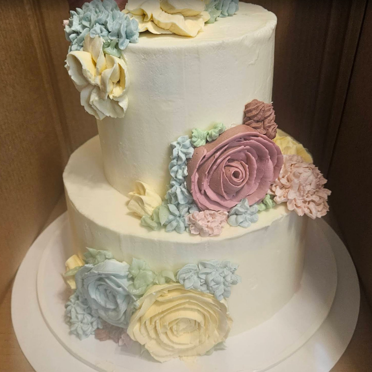 Two-tier ivory buttercream cake decorated with piped roses in blush, butter yellow and sage