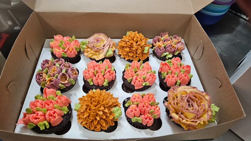 A dozen cupcakes in a bakery box, each piped with orange, coral and plum buttercream flowers
